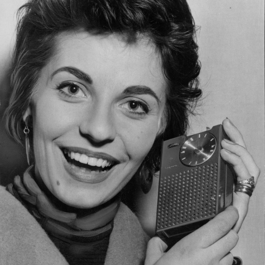 En kvinna lyssnar på årets nyhet 1955 - en rörlös transistorradio i västficksformat.